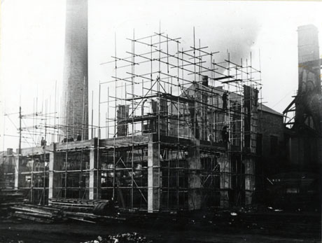 Photograph showing the concrete skeleton of the first and second floor of a building surrounded by scaffolding; four men can be seen in the distance standing on the first floor; behind the building is a large chimney, a high building and part of the winding gear of a colliery; the picture has been identified as South Hetton Colliery 1951, Building The Electric Winder House