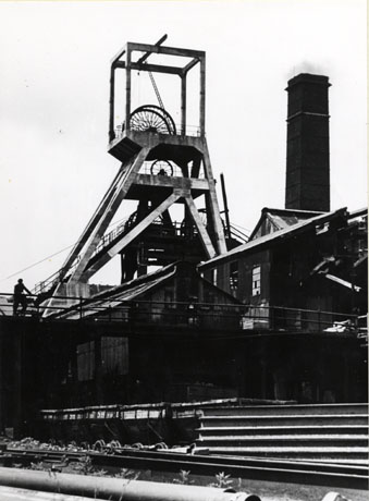 Photograph showing the winding gear of a colliery with further winding gear immediately behind it; in front of the winding gear are high corrugated iron buildings, a raised walkway, a line of coal waggons and piles of pipes and rails; the picture has been identified as depicting the new shaft head gear over the old shaft head gear at South Hetton Colliery