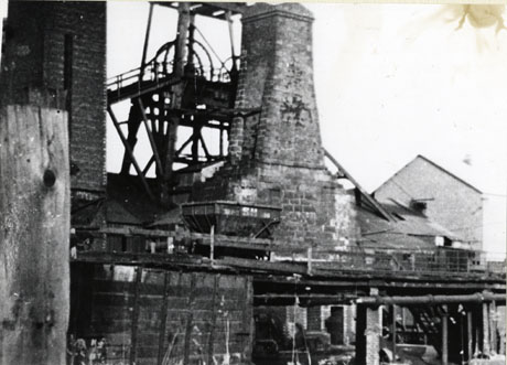 Photograph showing, in the foreground, a raised railway with a coal waggon with S H on its side; behind it, is a weathered brick chimney and, behind that, part of the winding gear; the colliery has been identified as South Hetton Colliery