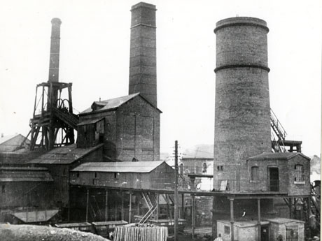 Photograph showing, on the left, the winding gear and, in the centre and on the right, a large square and a large round chimney of a colliery; in the foreground are small buildings, piles of logs, and two coal waggons; the colliery has been identified as South Hetton Colliery