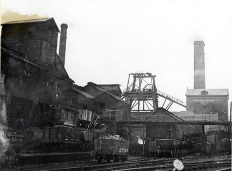 Photograph showing buildings of a colliery on the left, the winding gear in the middle, and a large chimney on the right, of the picture; in the foreground, are rail tracks and lines of coal waggons; the colliery has been identified as South Hetton Colliery