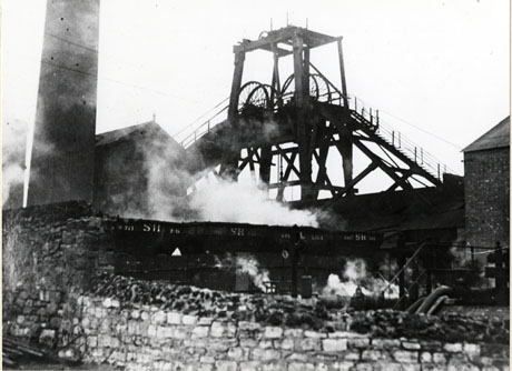 Photograph showing a wall in the foreground with steam rising immediately behind it; behind the steam are a chimney, coal waggons with S H on them, a building, and winding gear; the colliery has been identified as South Hetton Colliery