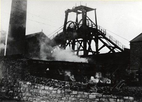 Photograph, taken from below, of the winding gear of a colliery, outlined against the sky; on the left of the picture are the bottom part of a tall chimney and the outline of the roof of a building; on the right is the outline of the side and roof of another building; in front of the winding gear can be seen, indistinctly, the sides of colliery wagons, bearing the letters S. H., and the side of a wall