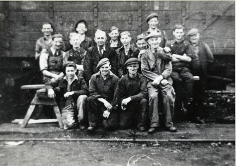 Photograph of sixteen men in overalls and caps, grouped in front of the side of a coal waggon, with a rail on the ground in front of them; they have been identified as a group of workers at South Hetton Colliery