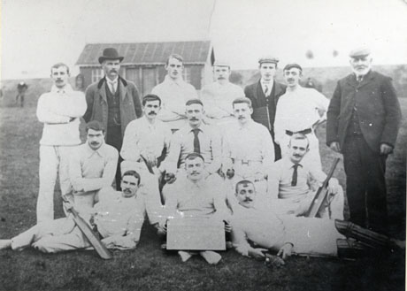 Photograph of twelve men in cricket whites, posed on a field with a small hut behind them in the distance; they are accompanied by a middle-aged man, a young man, and elderly man dressed in suits; a man on the front row is holding a notice, which cannot be read; they have been described as South Hetton Cricket Team, North Durham League Champions, 1901