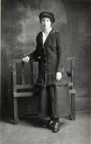 Photograph showing a woman in an ankle-length skirt, a long jacket with epaulettes, and a cap with the letters N. E. R. on it; she is leaning against a bench in a photographer's studio; she has been identified as Mrs. Wilson, porter at South Hetton Railway Station between 1914 and 1919