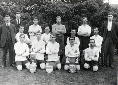 Photograph of twelve men in football strip, accompanied by three other men in suits, grouped in front of bushes; two large trophy cups and three shields containing medals are in front of the group; the group has been identified as members of South Hetton Royal Rovers Football Club