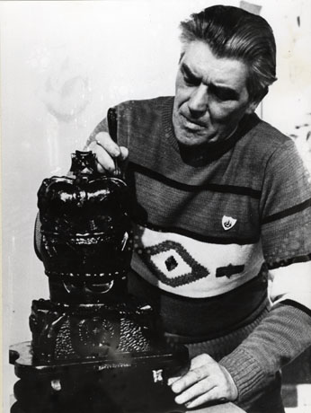 Photograph of the head and torso of a middle-aged man wearing a patterned jumper and what appears to be a Blue Peter badge; he has a tool in his right hand with which he is putting the finishing touches to a carving in coal of a crown which is on a stand in front of him; he has been identified as Dave Merrington of South Hetton with his triple crown carving