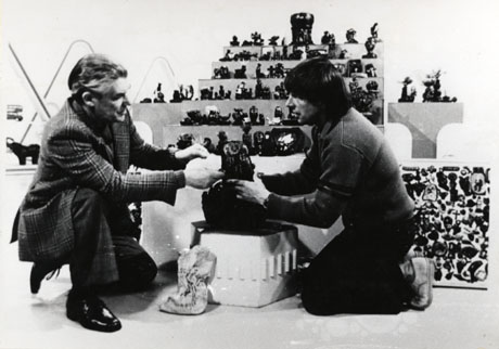 Photograph of Dave Merrington of South Hetton kneeling at the left of the picture showing one of his carvings in coal to John Noakes, a presenter on Blue Peter, a television programme for children; behind the two men are six shelves displaying Mr. Merrington's carvings in coal