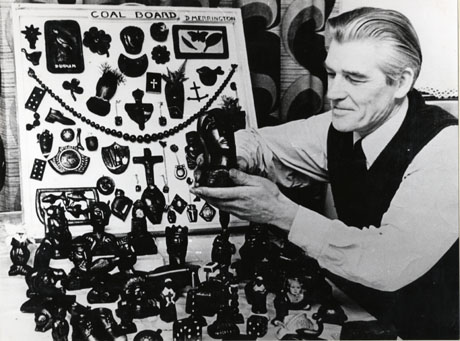 Photograph showing the head and torso of Dave Merrington of South Hetton, on the right of the picture, with a board displaying small carvings in coal and larger carvings in coal on a table in front of him