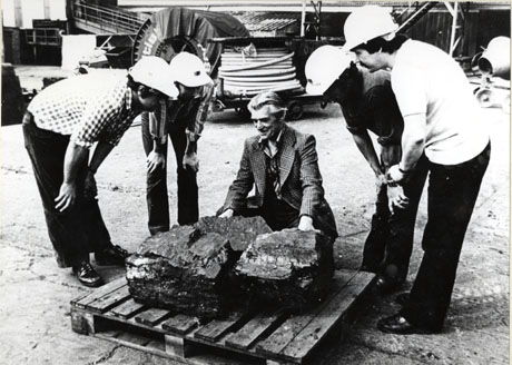 Photograph showing Dave Merrington of South Hetton in the centre of the picture, crouching behind a pallet on which there is a large lump of coal; four people in hard hats are standing on either side of the pallet; behind the group, coils of wire, a concrete mixer, low buildings, and steps can be seen; the photograph has been described as Dave Merrington of South Hetton selecting coal for carving at Haig Colliery, Whitehaven