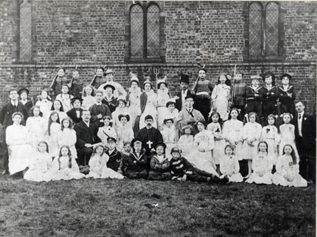 Photograph of approximately sixty men women and children in costume grouped against the wall of a church of brick with Gothic windows; the group has been identified as South Hetton Primitive Methodist Operetta Company; the members of the group are dressed as soldiers, sailors, clergymen, academics, gentlemen and Britannia