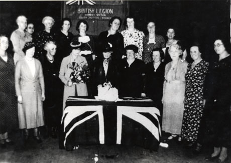 Photograph showing a group of nineteen middle-aged women standing round a table covered with a Union Jack, on which an iced cake is standing; one woman immediately behind the table is holding a bunch of flowers; behind the group is a board reading: British Legion Women's Section South Hetton Branch