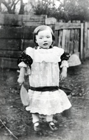 Photograph of a small boy, aged approximately three years, standing with a fence and trees behind him; he is dressed in a broderie anglaise pinafore with dark sleeves and a dark cummerbund; he is wearing button shoes and is holding a brush in his right hand; he has been identified as Amos Stubbs of South Hetton as a boy