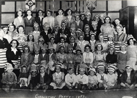 Photograph entitled Coronation Party- 1953, showing a group of approximately fifty children, aged between fourteen and four years, with eight adult women, posed against a wall with two shields on it; the children are wearing party hats; the party has been identified as taking place in South Hetton