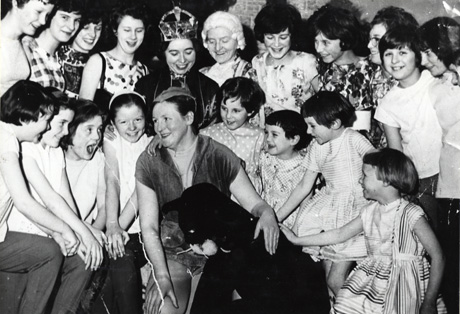 Photograph of seven young women and ten young girls, aged approximately between twelve and seven years, standing watching a woman dressed in a principal boy's costume, a woman dressed in a cloak and crown, and a stage cat; they have been identified as the members of the cast of the pantomime Dick Whittington staged by the South Hetton Womens' Institute