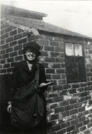 Photograph of a woman leaning against the wall of an outbuilding, wearing a hat with a brim, a jacket and carrying a large bag across the front of her body; in her left hand she is holding a number of letters; she has been identified as Mrs. E. Briggs, postwoman in South Hetton between 1939 and 1946, standing in the Eight Rows