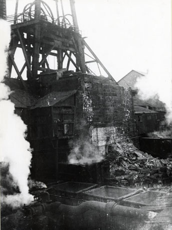Photograph showing part of the winding gear of a colliery with a brick pillar in front of it; in front of the pillar the interior of three coal trucks and a pipe giving off steam can be seen; the colliery has been identified as South Hetton Colliery