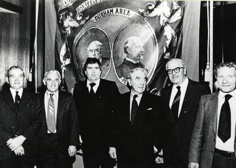 Photograph of part of the banner of South Hetton Lodge of the Durham Miners' Association, showing the portraits of Joe Gormley and Peter Lee, with six men standing in front of the banner; the man third from the right has been identified as Joe Gormley; the man second from the right as Lord Dormand; and the man first right as Arnold Ellis
