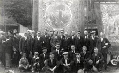 Photograph showing the banner of the South Hetton Lodge of the Durham Miners' Association displayed alongside the banner of Marley Hill Colliery Lodge; the banner has an unidentifiable scene with a female figure holding a flag and a wreath; in front of the banner, twenty three men and two boys are posed; behind the banner, trees can be seen; and to the left of the picture, five other men can be seen; the occasion has been identified as Durham Miners' Gala, 1932