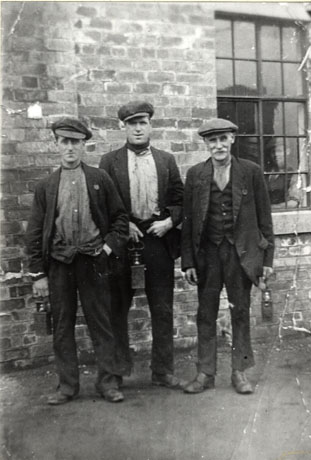 Photograph of three men in working suits and caps, carrying miners' lamps, standing against the wall and window of a brick building, identified as the Lamp Cabin, South Hetton Colliery
