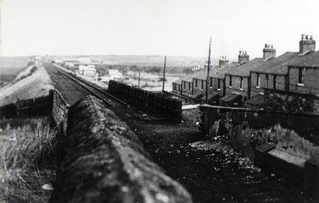 Photograph showing, in the middle of the picture, a railway line on an embankment running away from the camera; on the right of the picture, the first floors and roofs of a terrace of houses descending a slope can be seen; in the distance, there are indistinct buildings and fields; immediately in front of the camera is the top of a wall running away from the camera; the street has been identified as James street, South Hetton