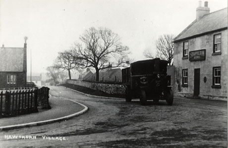 Postcard photograph, entitled Hawthorn Village, showing, on the left of the picture, the end of a building with a fence round it; in the middle of the picture a road runs away from the camera with a wall on its right-hand side, behind which the roofs of two houses and two trees can be seen; on the right of the picture, are the facade of The Stapylton Arms and a motor lorry in front of it, bearing the words South Hetton Coal Company
