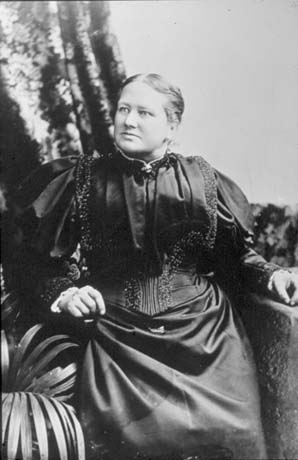 Photograph of a woman dressed in a blouse with mutton leg sleeves and elaborate decoration and a plain straight skirt in the same fabric; she is sitting on a chair with a piece of fabric behind her; she has been identified as Mrs. Kitchen of South Hetton