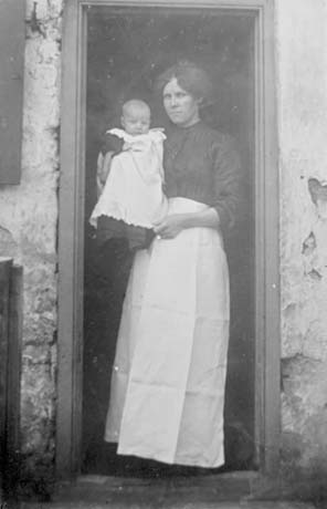 Photograph of woman dressed in a dark blouse and a long white apron, standing in the doorway of a house; she is carrying an infant in her arms, dressed in a white pinafore; they have been identified as Mrs. Rogers and William