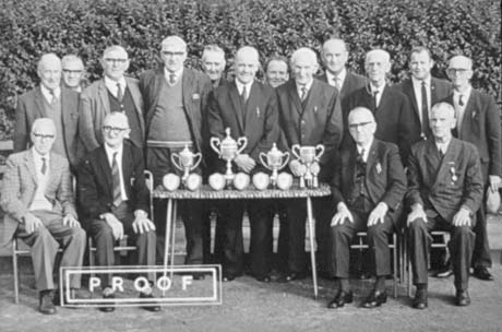 Photograph of a group of sixteen elderly men wearing suits and ties posed against a high hedge; in front of the group is a table bearing four trophy cups and six shields; the group has been identified as the Veteran Bowls Association of South Hetton at its presentation of trophies