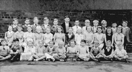Photograph of thirty five children, aged approximately five years, posed against a brick wall; a child on the front row; a child on the front row is holding a notice which reads South Hetton Infants Class 1 1949