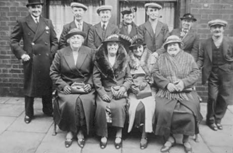 Photograph of a group of six elderly women and five elderly men, wearing overcoats and hats, posed against the wall and window of a building; they have been identified as a group from South Hetton on a day trip
