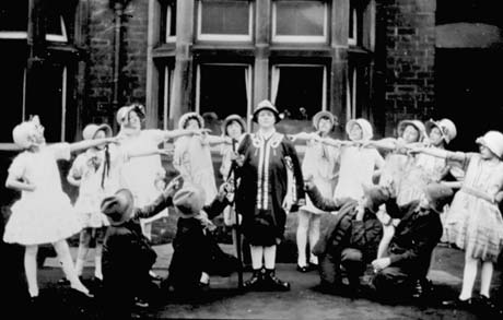 Photograph of ten girls, aged approximately between eight and twelve years, standing in a circle, dressed in short frilly frocks and bonnets, pointing at a figure standing in the middle of them; this woman is dressed in a Ruritanian uniform of cap, cloak, breeches and boots; in front of her are four people dressed in suits and hats, kneeling on the ground and pointing to the central figure; behind the group is a bay window; they have ben identified as taking part in School Operetta in South Hetton