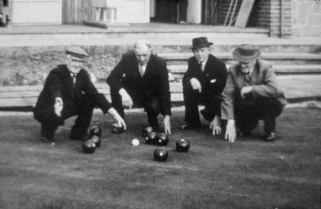 Photograph of four elderly men, wearing suits and ties, squatting on a bowling green; behind them are steps and part of the bottom of the walls of a building; in front of them are eight balls and a jack; they have been identified as being in South Hetton