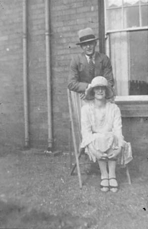 Photograph showing a woman, dressed in a summer frock and wearing a hat, sitting in a deckchair with a man in a suit and a Trilby hat, standing leaning on the back of the chair; behind the man and woman is the wall and window of a house; they have ben identified as Evelyn and James Lemmon of South Hetton