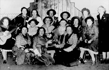 Photograph of fourteen middle-aged women, dressed as cowboys and cowgirls, grouped against the wall of a hall; one woman can be seen holding a guitar and another a ukelele; a woman wearing a formal suit is standing at the right of the picture; the group has been identified as Women's Institute Entertainers at South Hetton