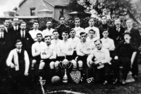 Photograph of fourteen men in football strip, accompanied by nine other men, posed against the high garden wall of a house, the top storey of which can be seen; in front of the group are a trophy cup, three shields bearing medals and a football; the group has been identified as the Royal Rovers, South Hetton