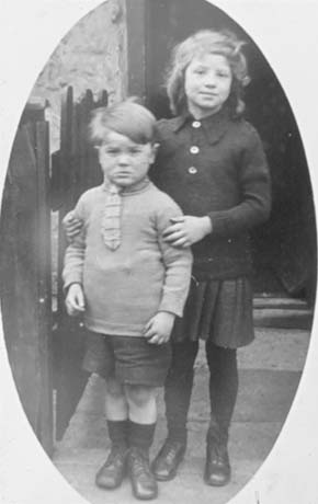Photograph showing a small boy, aged approximately six years, standing with a small girl, aged approximately eight years, near the fence of a house with its doorway behind them; the boy is wearing a jumper, tie, shorts, socks and boots; the girl is wearing a jumper with three large buttons, a pleated skirt, dark stockings and shoes; the have been identified as being in the Eight Rows, South Hetton
