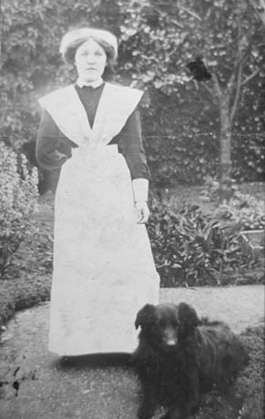 Photograph of a woman dressed in an apron down to her feet with a dark garment under it and large light cuffs; she is wearing a cap on her head; she is standing in a garden with a path and plants behind her; a dog is at her feet; she has been identified as Mrs. Haley in Service In East Rainton