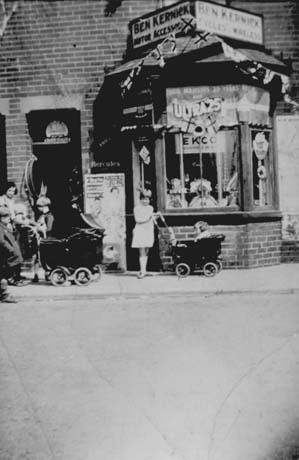 Photograph of the exterior of a shop with a bay window; above the window are signs reading as follows: Ben Kernick Motor Accessories and Ben Kernick Cycles and Wireless; in the window is a sign reading Their Majesties' 25 years and Ultra 25; it is, therefore, possible that the photograph was taken in 1935, the year of the Silver Jubilee of George V; a woman with three small children and a perambulator and a child with a perambulator are standing in front of the shop