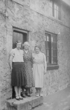 Photograph of a middle-aged woman wearing a patterned dress; a young woman wearing a blouse and skirt is on her right and a young woman wearing a sleeveless dress is on her left; all three are standing on the step of a stone house; they have been described as Mrs. Bell and Daughters in Hall Street, South Hetton