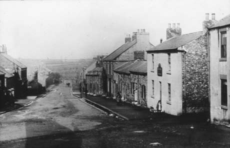 Photograph of a street running away from the camera downhill; in the distance fields can be seen; on the left of the picture the buildings are indistinct; on the right the facades of terraced houses and that of a public house can be seen; the street has been identified as Front Street, South Hetton; a small girl can be seen in front of the public house