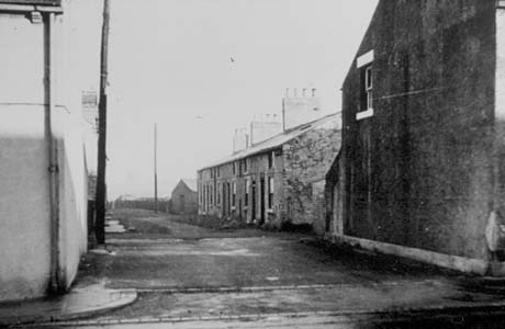 Photograph showing a back road running away from the camera with the sides of two houses at the front of the picture; in the distance the facade of a terrace of small cottages can be seen; the road has been identified as being in South Hetton