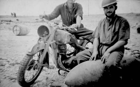 Photograph showing a motor bicycle with a man astride it; the face of the man astride the bicycle is not in shot; in front of the bicycle is a man sitting on a bomb; he is wearing a short-sleeved shirt, trousers and a tin hat and has been identified as Stoker Rackstraw of South Hetton, at Tobruk, North Africa, presumably during the Second World War; behind the men, a flat terrain with small indistinct figures in the distance can be seen