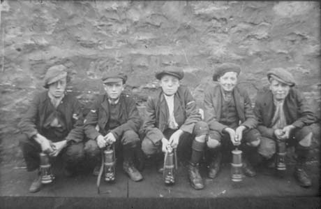 Photograph showing five boys aged approximately fourteen years squatting with their backs to a wall; each is wearing a cap, boots, jacket, waistcoat, long socks, and is carrying a miner's lamp; they have been described as Five Pit Lads at South Hetton