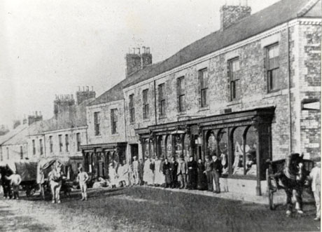 Photograph showing the facades of large terraces with shop windows on the ground floor on the right of the picture; in front of the shop windows are sixteen people lined up; they cannot be identified; two covered waggons with horses hitched to them can be seen in the road with a man standing near them; the photograph has been identified as Station Town Co-operative Premises, 1894