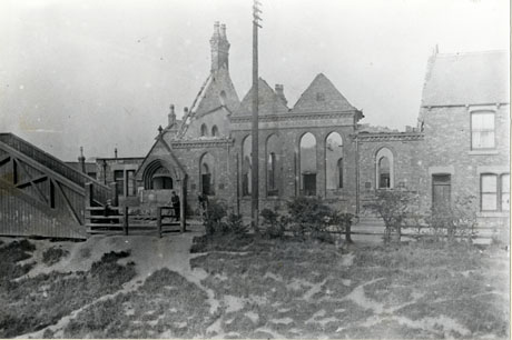 Photograph showing the side walls, the porch, and the end and back walls of a church, left standing after a fire; the church has the words Primitive Methodist Church and School 1893on it; at the left of the picture, the end of a footbridge can be seen; the church adjoins a terraced house on the right of the picture; a road and a piece of grass are in the foreground; the photograph has been identified as depicting Station Town