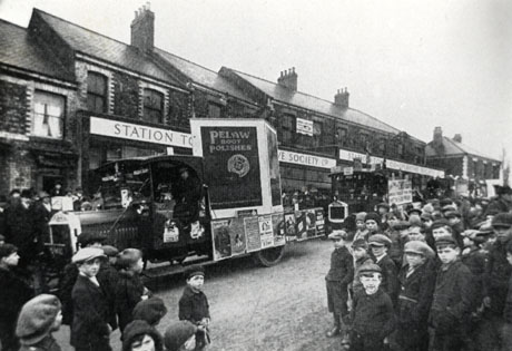 Photograph showing, at the back of the picture, the facade of Station Town Co-operative Society shops; in front of the shops, are three motor lorries with advertisements on them; the first lorry carries a large advertisement for Pelaw Boot Polishes; the other advertisements cannot be read; crowds of people can be seen on both sides of the road in front of the shops; the road has been identified as Station Lane