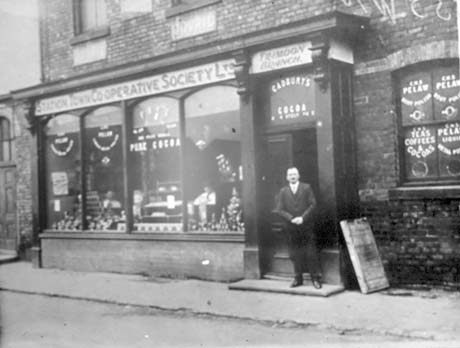 Photograph showing the exterior of the Trimdon Branch of Station Town Co-operative Society; the windows of the shop can be seen advertising Cadbury's Cocoa and the Co-operative Wholesale Society Pelaw Boot Polish and Co-operative Wholesale Society Teas Coffes and Cocoas; a man dressed in a dark suit is standing on the doorstep of the shop