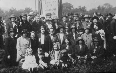 Photograph of a group of approximately fifty women, dressed in overcoats and suits, and four children, aged between three and seven years, posed in a field; behind them is a banner reading Station Town Women's Labour..